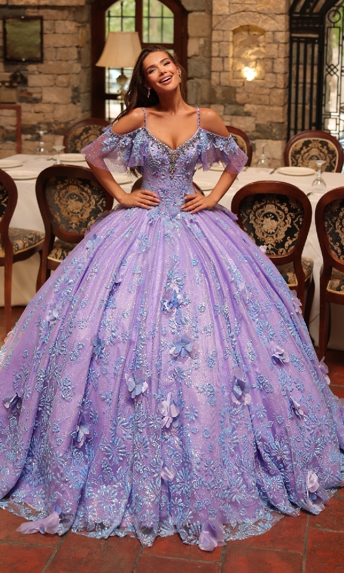 A woman stands indoors wearing the Amarra 54229 Glitter Tulle Quinceañera Dress, a floor-length, off-shoulder lavender ball gown adorned with floral embroidery, delicate beading, and sparkling glitter tulle embellishments.